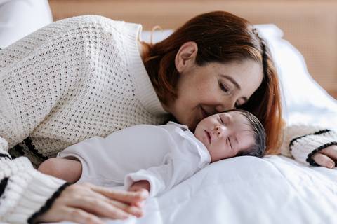 Foto da mãe deitada de lado na cama, de bochecha colada com a bebê em momento de aconchego.'