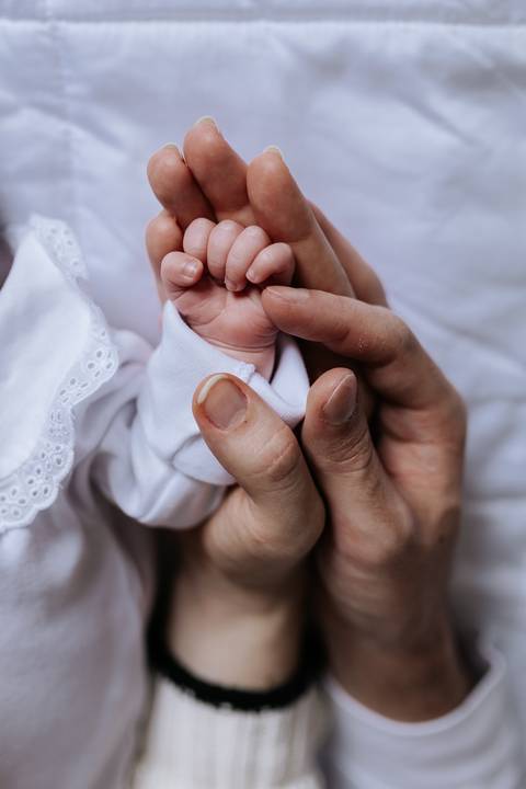 Detalhe macro das mãos dos pais envolvendo com cuidado os dedinhos minúsculos da recém-nascida.'