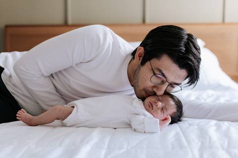 Foto do pai deitado na cama dando um beijo carinhoso na cabeça da bebê Nely.'