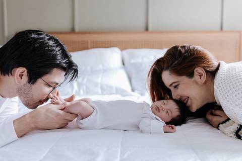 Foto do casal deitado na cama sorrindo e admirando a bebê deitada entre eles.'