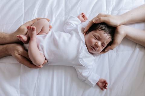 Foto de ângulo superior da bebê Nely na cama, amparada simetricamente pelas mãos dos pais na cabeça e nos pés.'