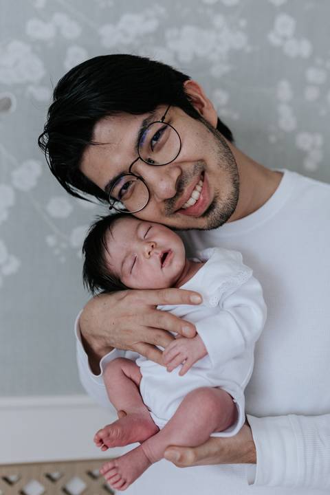 Close do pai sorridente e de óculos, aconchegando a bebê Nely junto ao seu rosto.'