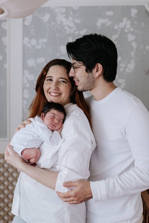 Retrato oficial do casal sorridente com a bebê Nely adormecida no aconchego do colo da mãe.'