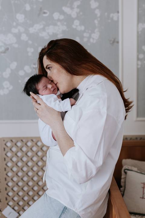 Foto de perfil da mãe em pé, dando um beijo carinhoso na testa da bebê Nely no colo.'