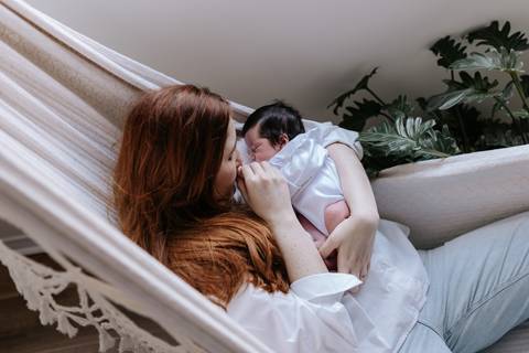 Foto da mãe deitada em uma rede de descanso branca, aninhando a bebê junto ao peito.'