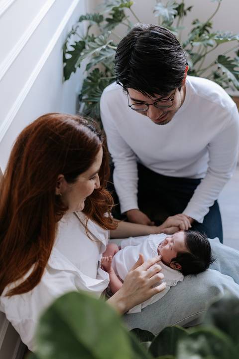 Foto do casal inclinado no chão cuidando e sorrindo para a recém-nascida deitada à frente deles.'