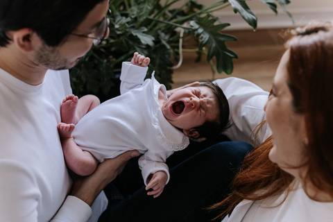 Foto divertida da bebê Nely dando um bocejo bem grande no colo do pai, com a mãe sorrindo ao lado.'