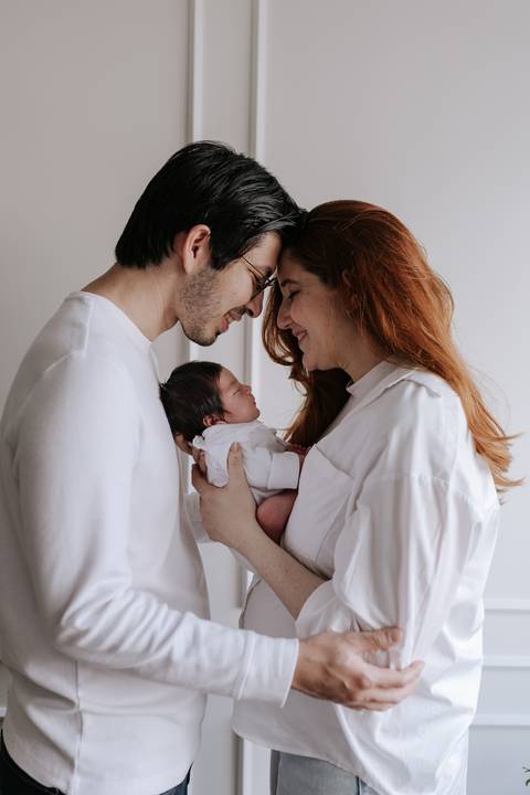Retrato de perfil do casal encostando as testas com carinho enquanto a mãe segura a bebê nos braços.'