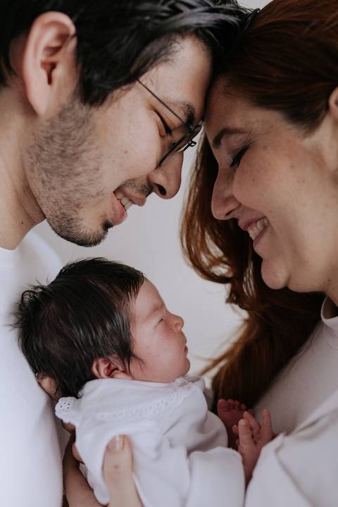 Retrato de close em perfil do casal sorrindo um para o outro com a bebê Nely entre eles.'