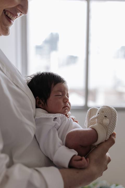 Foto aproximada da bebê dormindo no colo, destacando sua touquinha e sapatinho de tricô claro.'