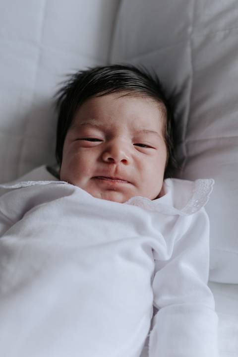 Close-up da bebê Nely com os olhinhos abertos e uma expressão expressiva no rosto, deitada em um fundo claro.'