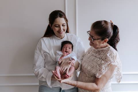 Retrato carinhoso de três gerações reunidas, mostrando a mãe e a avó segurando juntas a bebê Nely no colo.'