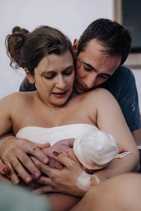Retrato do casal olhando fixamente para o bebê aninhado no colo materno, logo após o parto.'