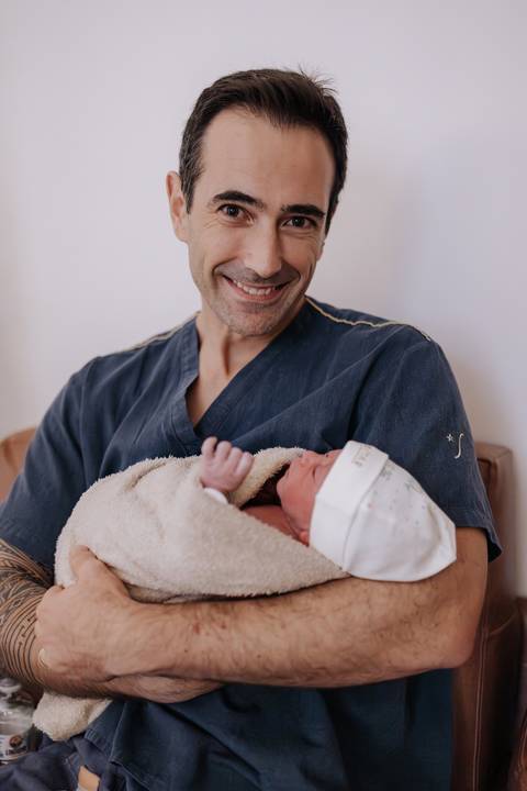 Retrato do pai sorridente segurando o filho no colo, envolto em uma manta bege.'