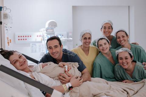 Retrato oficial com o casal no leito e a bebê, cercados pelas profissionais da equipe médica sorridentes.'