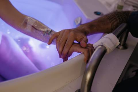 Detalhe das mãos do casal firmemente entrelaçadas na borda da banheira de parto com água iluminada.'