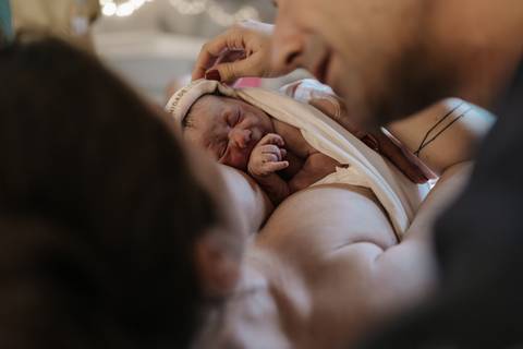 Foto aproximada do bebê recém-nascido chorando no peito, sob os olhares maravilhados dos pais.'