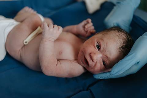 Foto do bebê deitado de costas na mesa de exames recebendo os primeiros cuidados da equipe médica.'