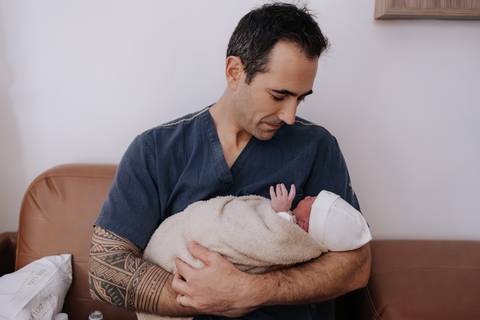Foto do pai sentado no sofá da maternidade, olhando com ternura para o bebê recém-nascido aconchegado em seus braços.'