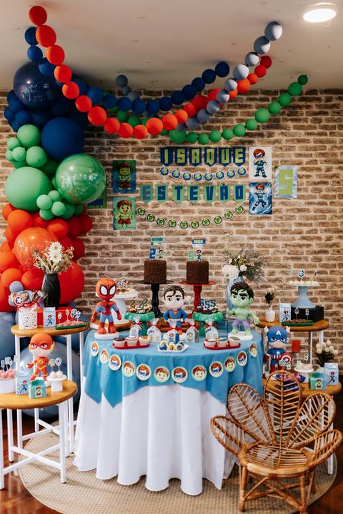 Foto panorâmica da mesa de doces principal decorada com balões coloridos e fundo imitando parede de tijolos.'