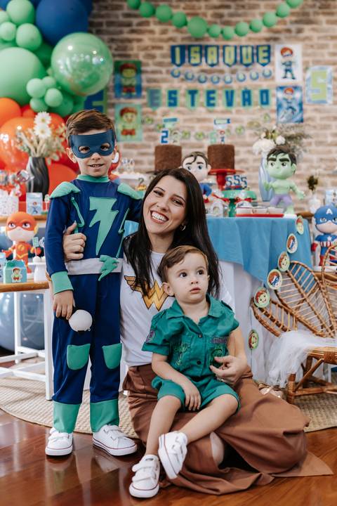 Retrato da mãe sorridente com os dois filhos, com o mais velho usando fantasia de super-herói azul e máscara.'