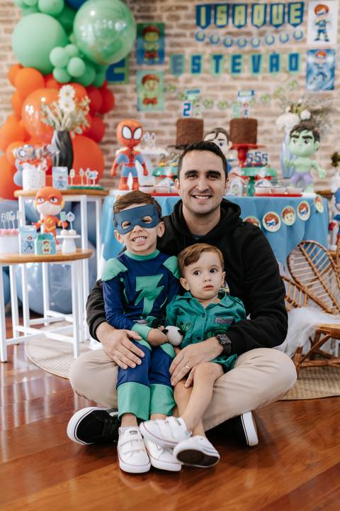 Retrato do pai abraçado com os dois filhos no chão em frente à mesa do bolo.'