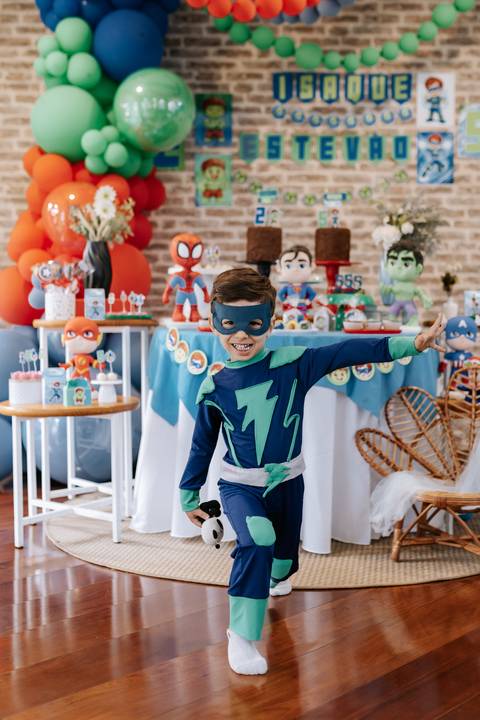Foto divertida do aniversariante fantasiado de herói fazendo pose com os braços abertos ao lado da decoração.'