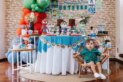 Foto do caçula sentado em uma mini cadeira de vime ao lado da mesa de doces principal.'