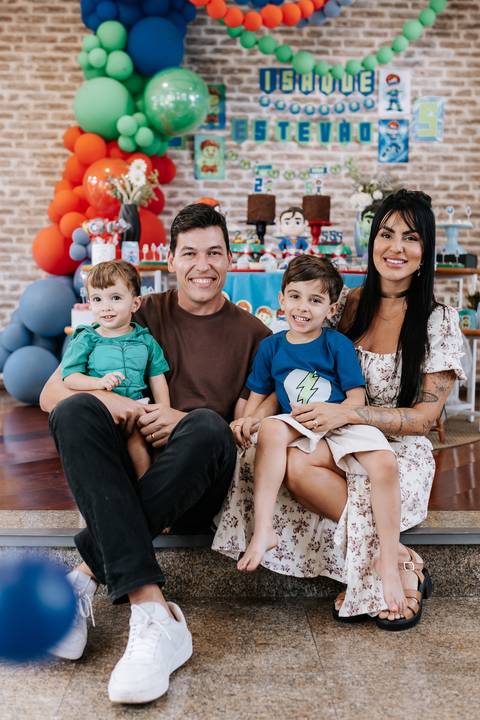 Retrato da família reunida no chão ao lado de convidados na frente do painel da festa.'