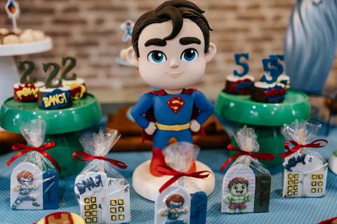 Detalhe da mesa do bolo com um boneco decorativo do Super-Homem cercado por docinhos e sacolinhas surpresa.'