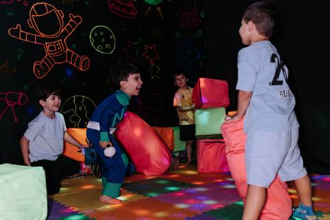 Foto das crianças brincando com blocos de espuma gigantes na pista de dança iluminada da festa.'