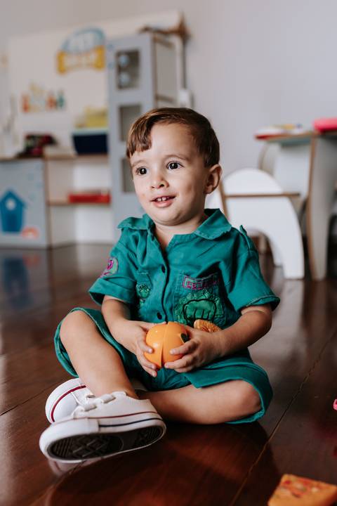 Registro espontâneo do caçula sentado no chão segurando um brinquedo laranja.'