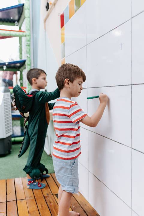Registro de crianças concentradas desenhando com canetinha em um painel branco de azulejos.'