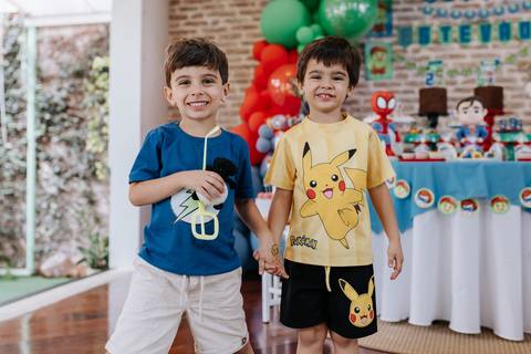Retrato de dois amiguinhos sorridentes, um deles com camiseta do Pikachu, posando juntos perto da mesa do bolo.'