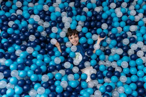 Registro divertido do aniversariante sorrindo submerso em meio a uma piscina de bolinhas azuis e brancas.'