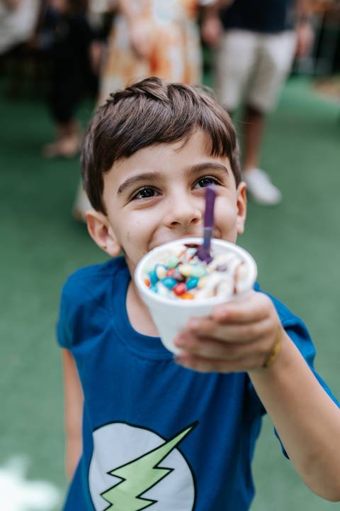 Close-up de um dos meninos sorrindo largo enquanto segura um copo de sorvete cheio de confeitos coloridos.'