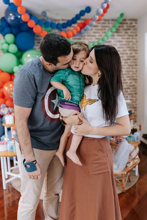 Retrato aproximado do casal trocando carinhos com o filho caçula no colo em frente à decoração.'