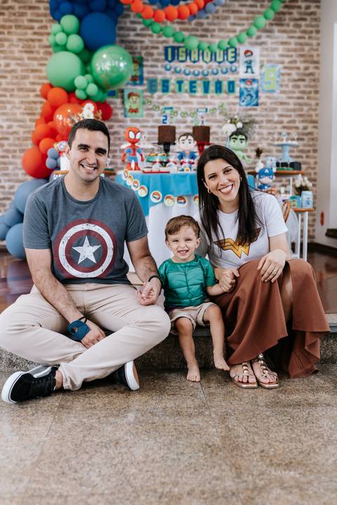 Retrato do casal sorridente sentado no chão da festa ao lado do filho menor.'