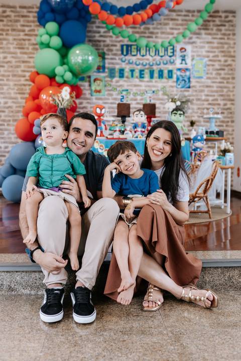Retrato oficial com a família completa reunida e sorridente em frente à mesa principal de super-heróis.'