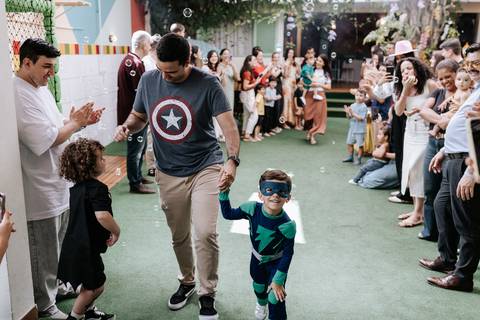 Foto em ângulo aberto mostrando o pai e o filho fantasiado caminhando entre os convidados que aplaudem.'