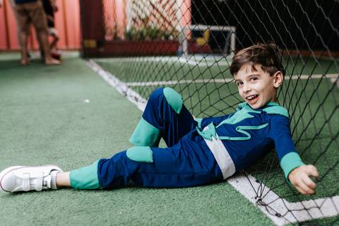 Foto do aniversariante mais velho deitado de lado no gramado sintético da quadra de futebol do buffet, sorrindo para a câmera.'