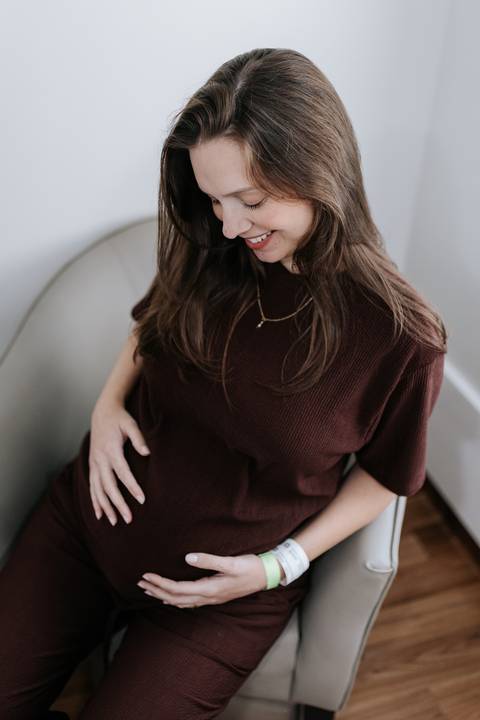 Foto de ângulo superior da gestante sorridente com blusa marrom, acariciando a barriga no hospital.'