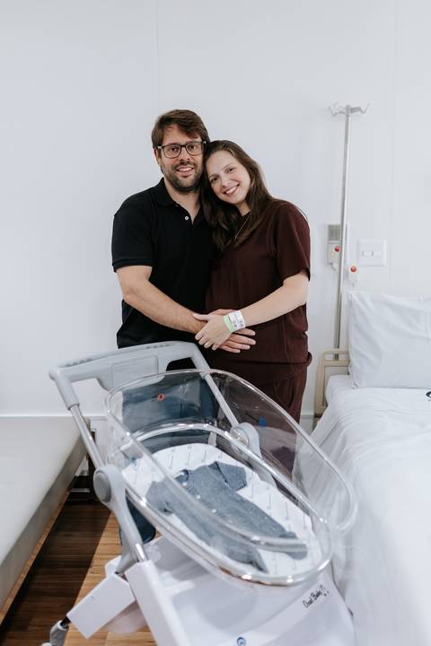 Retrato do casal posando sorridente em pé atrás do berço de acrílico vazio no quarto da maternidade.'
