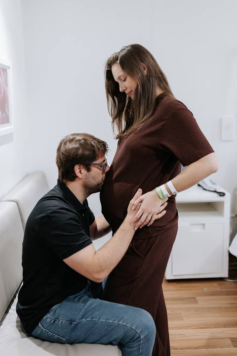 Foto de perfil do pai ajoelhado dando um beijo terno na barriga da gestante no quarto do hospital.'