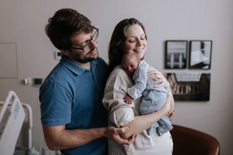Retrato do casal no quarto do hospital, com a mãe segurando o bebê e o pai observando orgulhoso ao lado.'