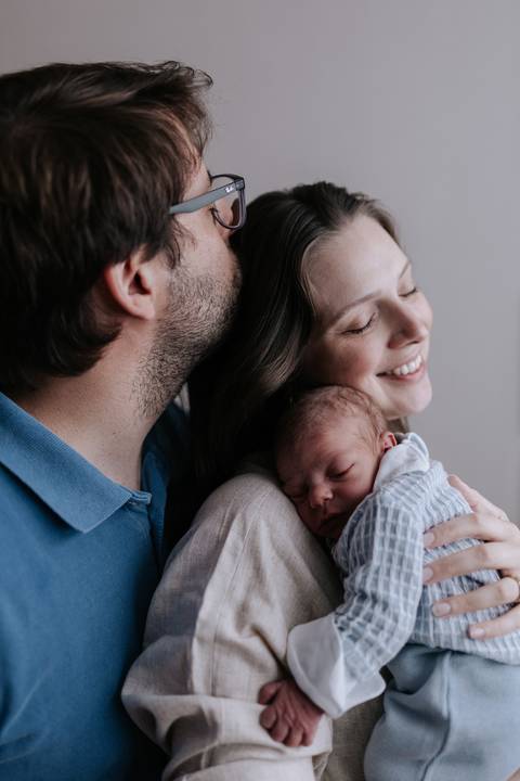 Close do pai dando um beijo carinhoso no topo da cabeça da mãe enquanto ela segura o filho no colo.'