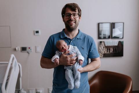 Retrato do pai em pé no quarto da maternidade, segurando o bebê Pedro com as duas mãos junto ao peito.'