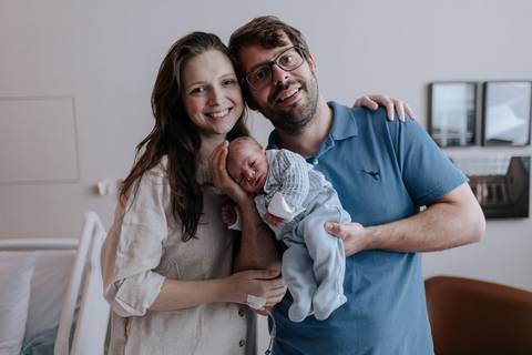 Retrato oficial do casal sorridente e abraçado, com o bebê Pedro no colo em seu primeiro dia de vida.'