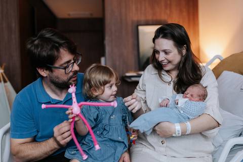 Retrato dos pais com os dois filhos no colo; o pai interage com a menina usando um brinquedo rosa.'