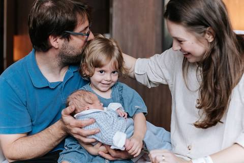irmã mais velha pegando o mais novo no colo, pai e mãe felizes ao lado. '